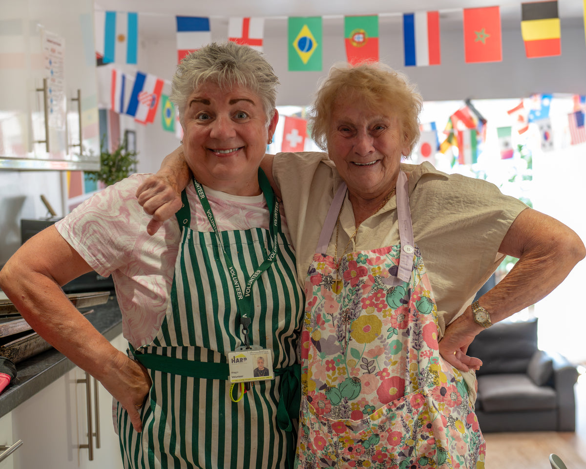 Two smiling women standing side by side in a brightly lit kitchen or cafe, each wearing an apron. The woman on the left wears a green and white striped apron over a light pink shirt, and the woman on the right wears a colorful floral print apron over a beige top.