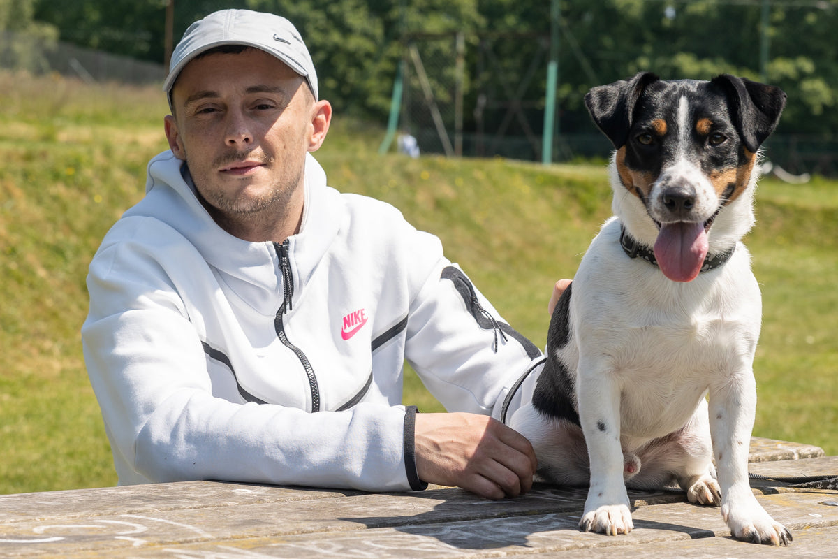 A man wearing a white Nike zip-up hoodie and a white cap sits outdoors next to a black, white, and tan dog on a stone table in a grassy park.